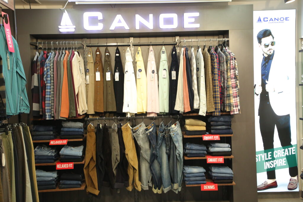 A premium menswear retail display featuring neatly arranged shirts, jackets, trousers, and denim jeans under the illuminated "CANOE" brand sign. The display showcases a variety of casual and formal clothing in different colors, with folded jeans and trousers organized on wooden shelves inside a modern clothing store.