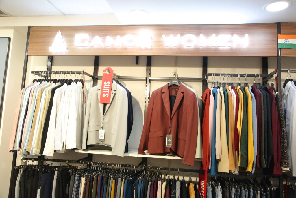 A modern women's fashion section in a retail clothing store featuring a stylish collection of blazers, shirts, trousers, sweaters, and tops displayed on organized racks beneath an illuminated "CANOE WOMEN" sign. The display showcases a variety of colors and contemporary designs in a clean, premium shopping environment.
