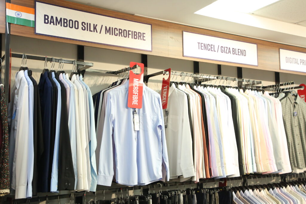 A premium retail clothing display featuring a collection of formal men's shirts organized by fabric categories, including Bamboo Silk/Microfibre, Tencel/Giza Blend, and Digital Print. The shirts are neatly arranged on racks in a modern showroom, showcasing a variety of classic colors, pastel shades, and premium fabrics.