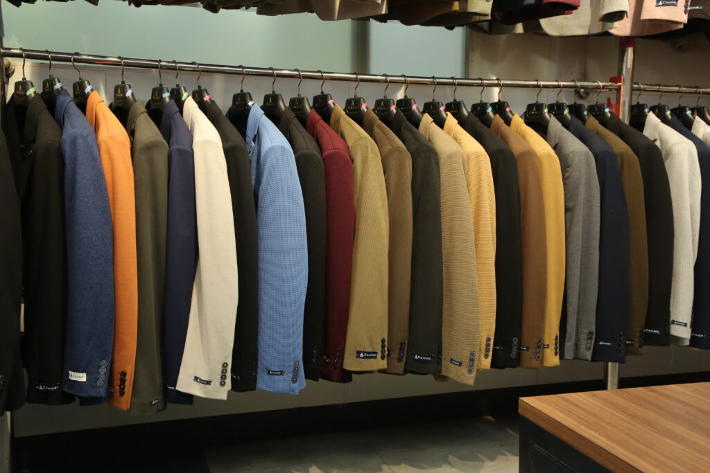 A premium collection of men's blazers displayed on a clothing rack in a modern fashion store. The blazers are available in a wide range of colors, including black, navy, white, beige, maroon, blue, gray, olive, and mustard, showcasing elegant tailoring for formal, business, and smart-casual occasions.